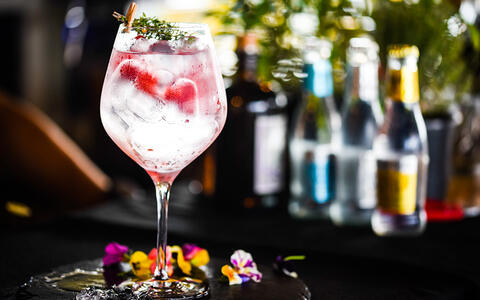 Cocktailglas mit Drink "Berlin Berry", Blütendekor und Tonic Water im Hintergrund